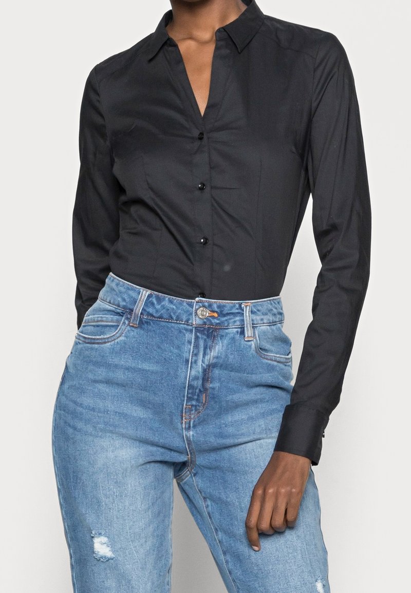 Personne portant une chemise noire ajustée à boutons avec manches longues et un jean taille haute bleu clair avec une légère usure.