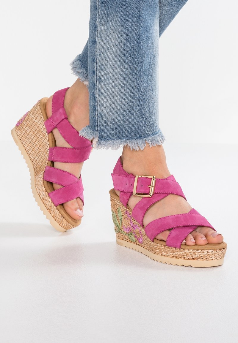 Pieds portant des sandales compensées roses à brides avec broderies florales sur les côtés, associées à un jean bleu à ourlet effiloché.