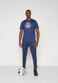Mørkeblå Nike t-shirt med Paris Saint-Germain-logo, parret med matchende mørkeblå træningsbukser og sorte sportsko, ved siden af en mønstret fodbold.