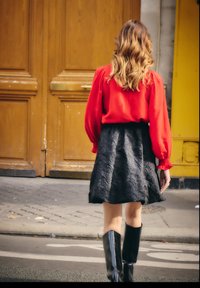 Frau mit welligem Haar, die eine rote Bluse, einen schwarzen Blumenrock und schwarze Stiefel trägt und auf einem Bürgersteig in der Stadt in der Nähe von Holztüren geht.