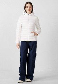 Witte gewatteerde jas met capuchon en rits aan de voorkant, met horizontale stiknaden. Gecombineerd met marineblauwe cargobroek met zijzakken en wijde pijpen.