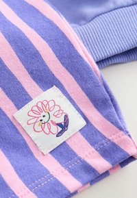 Tessuto rigato viola e rosa con un'etichetta cucita che presenta un design floreale. Morbida texture, stile informale e dettaglio a coste blu sui polsini.