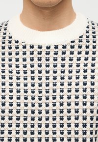 Homme portant un pull en tricot blanc avec des motifs noirs verticaux en pointillés et un col ras du cou épais côtelé, sur un fond uni.