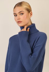 Niebieski sweter dziany z wysokim, prążkowanym kołnierzem i długimi rękawami, o luźnym kroju i miękkiej fakturze.