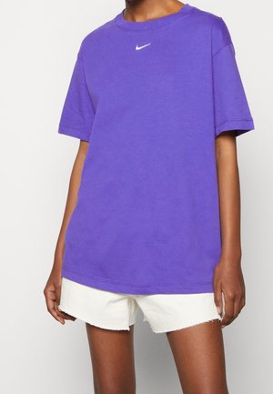 Personne portant un t-shirt Nike violet ample et un short blanc effiloché, debout devant un fond clair uni.