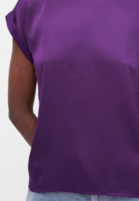 Άτομο που φοράει μπλούζα μωβ με κοντά μανίκια με γυρισμένα μανσέτα και ανοιχτόχρωμο τζιν, με χαλαρά χέρια πίσω από την πλάτη.