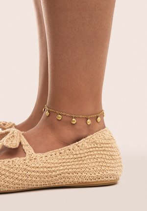 Caviglia con bracciale in oro a catena con piccoli charm a forma di conchiglia, abbinata a scarpe piatte beige all'uncinetto su uno sfondo neutro.