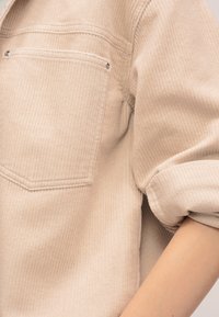 Μπεζ πουκάμισο από κοτλέ με εμπρόσθιο τσέπη στο στήθος και αναδιπλωμένο μανίκι που αποκαλύπτει μέρος του βραχίονα.