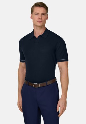 Mann trägt ein marineblaues Poloshirt mit weißem Ärmelsaum, braunen Gürtel und marineblaue Hose, steht vor einem schlichten hellen Hintergrund.