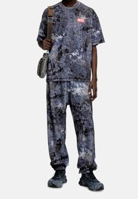 Muž oblečený v odpovídajícím modro-černém tričku se abstraktním vzorem a teplácích, v černých robustních teniskách a s křížovou taškou.