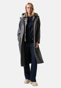 Grijze lang model jas van een wolmix met een zilverkleurige gewatteerde binnenlaag, capuchon, brede mouwen, gecombineerd met een marineblauw shirt en wijde spijkerbroeken met uitlopende pijpen. Crèmekleurige chunky sneakers.