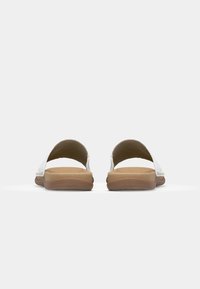 Sandales blanches à semelle plate avec une tige en cuir lisse, arrière ouvert et semelles en caoutchouc tan. Présente une couture minimale le long des bords.