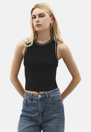 Jonge vrouw met blond haar, gekleed in een zwarte mouwloze cropped top met gouden biezen en een hoge taille blauwe spijkerbroek, poserend met de handen achter de rug.