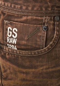Vaqueros de mezclilla marrón con un acabado texturizado, que presentan una etiqueta en el bolsillo trasero que dice "GS RAW 5204" y detalles de costura en contraste.