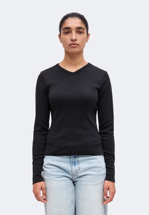 Femme aux cheveux foncés tirés en arrière, portant un haut noir à manches longues avec un col en V et un jean bleu clair sur un fond clair uni.
