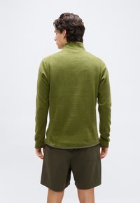 Άνδρας με πλάτη φορά υφασμάτινη φλις μπλούζα με μακριά μανίκια σε χρώμα ελιάς και σκούρο πράσινο σορτς, σε απλό λευκό φόντο.