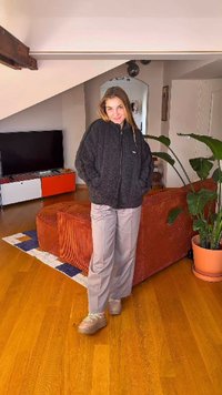 Zwarte fleecevest met ritssluiting, lichtgrijze wijde broek, beige sneakers met plateauzool. Kamer met houten vloer, oranje bank en groene plant.