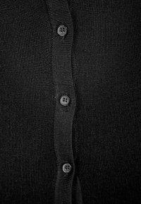 Černý pletený cardigan s texturovaným povrchem, zapínání na knoflíky a šesti kulatými černými knoflíky rovnoměrně rozmístěnými vpředu.