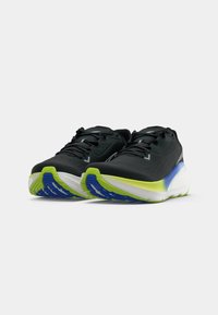 Baskets de sport noires avec un dessus texturé, présentant une semelle intermédiaire blanche et des accents vifs en vert et bleu sur la semelle extérieure. Design à lacets.
