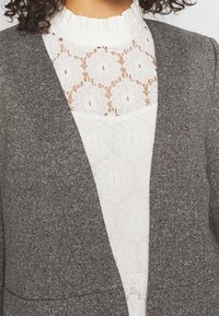 Blazer en maille grise à devant ouvert, associé à un haut en dentelle blanche avec motifs floraux et col à volants. Look texturé et superposé.