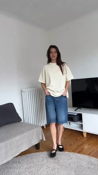 Moteris stovi svetainėje, dėvi laisvą smėlio spalvos marškinėlį, kelio ilgio džinsinius šortus ir juodus Mary Jane batelius, rankos kišenėse.