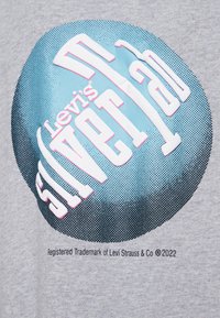 Camiseta gris con un gráfico redondo azul y blanco que presenta "Levi's SilverTab" en texto en negrita. Patrón de puntos y etiqueta de marca debajo.