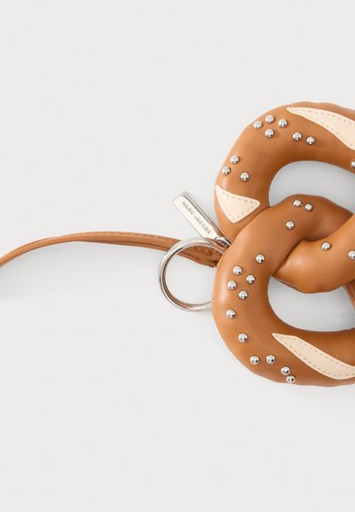 Porte-clés en cuir marron en forme de bretzel avec des clous argentés, des accents en cuir crème, un anneau métallique et une fine lanière en cuir sur fond blanc.