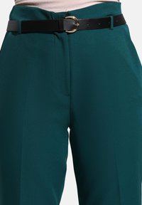 Pantalons taille haute en teal avec un pli avant, associés à une ceinture en cuir noir avec une boucle dorée ronde et une légère brillance.