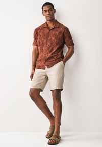 Chemise à manches courtes de couleur brun rougeâtre avec motif, short en lin beige, et sandales marron. La chemise présente un design texturé avec des formes géométriques.