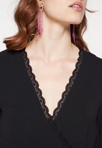 Haut cache-cœur noir avec un col en V orné d'un bord en dentelle festonnée. Détails remarquables : une texture lisse et une coupe ajustée. Boucles d'oreilles pendantes roses scintillantes.