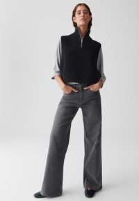 Frau steht mit den Händen in den Taschen, trägt eine schwarze ärmellose Zip-Up-Weste, ein graues Langarmshirt, weite, dunkle Jeans und schwarze Schuhe.