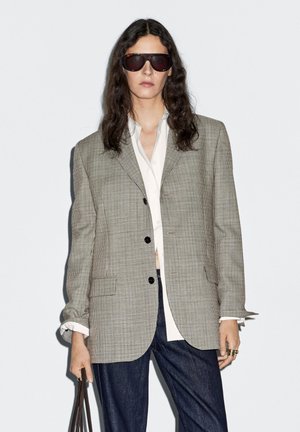 Blazer oversize a quadri marroni e bianchi realizzato in tessuto morbido, con bottoni neri e due tasche frontali, abbinato a una camicia bianca e jeans scuri.