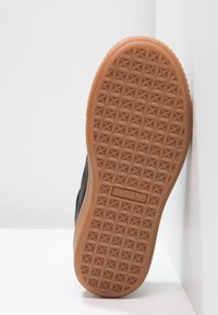 Semelle en caoutchouc marron d'une basket avec un motif triangulaire et le logo Puma, reposant contre un mur blanc.