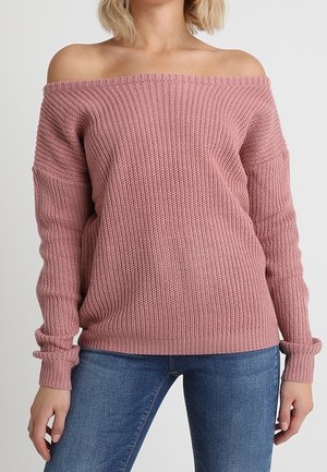 Femme portant un pull en tricot rose avec les épaules dénudées et un jean bleu, debout devant un fond clair uni.