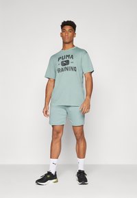 Set de tricou de antrenament și pantaloni scurți mint green, cu un grafic al logo-ului "PUMA" în negru. Modelul poartă încălțăminte sport neagră și șosete albe.