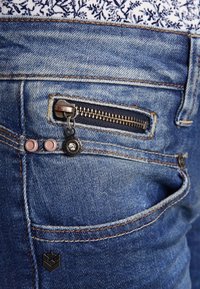 Denimjeans med en strukturerad blåtvätt, som har en liten dragkedjeficka, dekorativ hårdvara och kontrasterande sömmar.