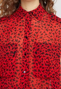Červená průhledná košile s černým leopardím potiskem, klasickým límcem, předními klapkami na kapsách a černými knoflíkovými uzávěry.