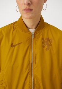 Jachetă bomber galbenă, fabricată dintr-un material ușor și neted. Caracteristici: închidere cu fermoar în față, guler ribb și logo Nike cu un design de leu.