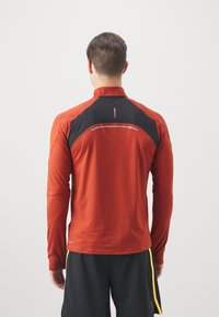 Langärmliges Sporthemd in Rot mit schwarzen Akzenten, ausgestattet mit einem hohen Kragen und einem reflektierenden Streifen auf dem Rücken für Sichtbarkeit. Glatte Textur.