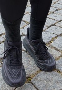 Scarpe sportive nere con tomaia testurizzata, design con lacci e colletto imbottito, abbinate a calzini a coste scuri, posizionate su una superficie di ciottoli.