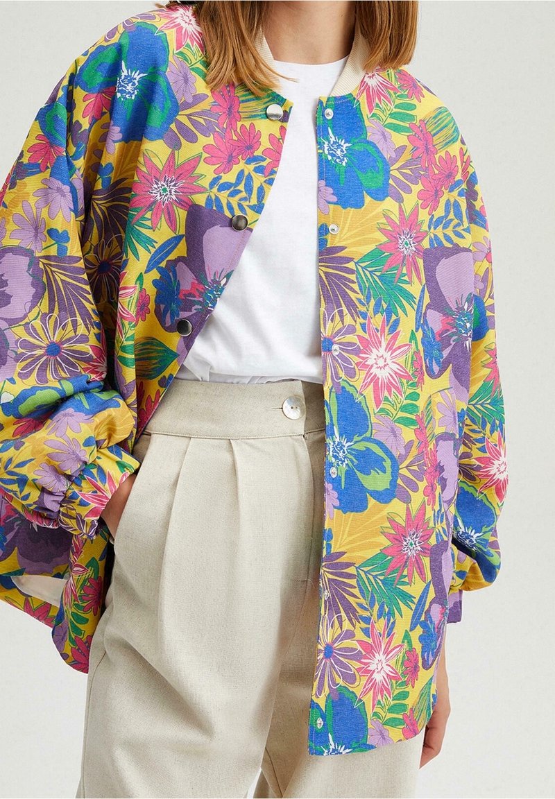 Kobieta w beżowych spodniach z wysokim stanem, białej koszuli oraz luźnej żółtej kurtce w dużą niebieską, różową i fioletową kwiecistą wzór.