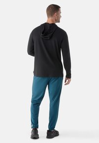 Schwarzes Kapuzenshirt mit langen Ärmeln, kombiniert mit türkisfarbenen, elastischen Hosen, die einen entspannten Schnitt und eine glatte Textur bieten. Schwarze Schuhe runden den Look ab.