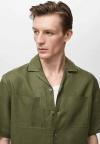 Short-sleeve olive green shirt made of linen, featuring two front pockets, a button closure, and a collar. Smooth texture and lightweight design.