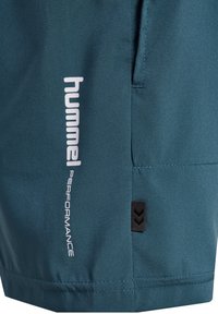 Teal idrottsshorts tillverkade av lättviktigt material. Har en vit "hummel PERFORMANCE"-logotyp och en svart etikett med chevrondetaljer.
