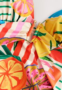 Gros plan sur un tissu coloré avec des imprimés de fruits et de fleurs, rassemblé et relié par un anneau en or avec des enveloppes en tissu corail et jaune.