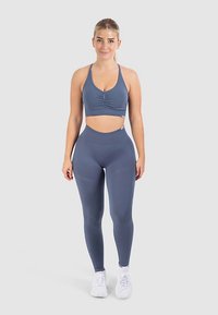 Grijze sportbeha met plooien aan de voorkant, gecombineerd met een high-waist legging in dezelfde kleur. Het materiaal lijkt soepel, rekbaar en aansluitend te zijn.