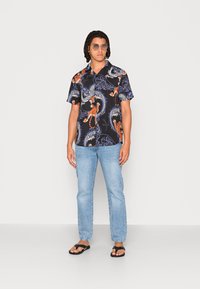 Kortærmet skjorte med knapper og sort baggrund, der har orange tigre og blå drager, parret med lyseblå jeans og sorte flip-flops.