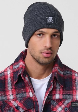 Gorro de punto acanalado gris oscuro con un parche de logo. El modelo lleva una camisa de franela a cuadros rojos y azules sobre una camiseta blanca.