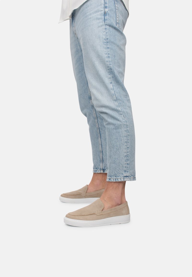 Scarpe slip-on beige in materiali scamosciati con suola in gomma bianca, abbinate a jeans azzurri con risvolto. Le scarpe presentano dettagli di cucitura minimi.