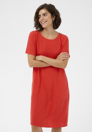 Femme aux cheveux courts et bouclés portant une robe rouge jusqu'aux genoux, souriant et regardant sur le côté, les bras croisés devant un fond uni.